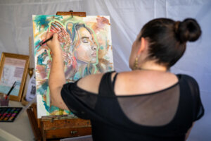 A woman with dark hair in a bun paints a colorful portrait of a woman with feathers on an easel. Art supplies and framed information sit nearby on the table.