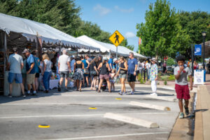 People browse under white tents at an outdoor market on a sunny day. A boy in a red shirt walks while eating ice cream in the foreground. Green trees and a pedestrian crossing sign are visible.