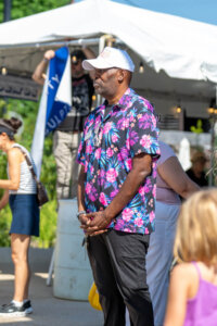 A man wearing a white cap and a vibrant floral shirt stands outdoors at an event, with people and a white tent visible in the background on a sunny day.