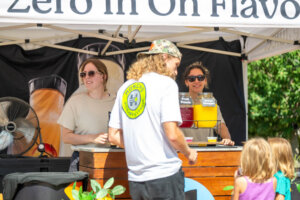 Two women stand behind a juice stand with jars of fruit punch and orange juice. A man in a white shirt with a logo and three children are in front of the stand, outdoors on a sunny day.