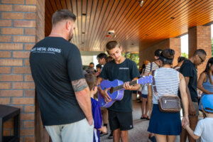 A boy in a black shirt plays a purple guitar in a crowd under a wooden ceiling. People around him, including a man in a black shirt and a woman with a bun, watch and interact.