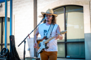 A man with long curly hair, wearing a hat and sunglasses, plays an electric guitar and sings into a microphone outside a building on a sunny day.