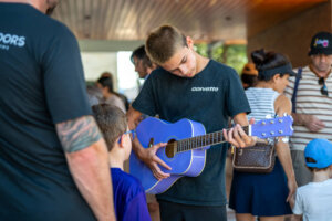 A boy plays a purple acoustic guitar while others, including children and adults, gather around him indoors. The scene appears casual and social, with people engaged and observing.