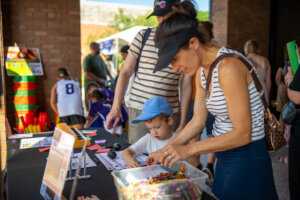 A woman and young boy do arts and crafts at an outdoor table with supplies. Other adults and children are gathered nearby. The event appears to be family-friendly and interactive.