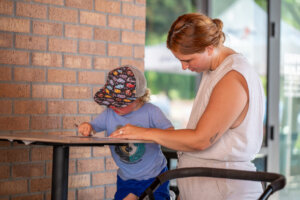 A woman helps a young child in a cap draw on paper at a tall table inside a building with brick walls and glass doors.