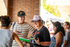 A group of people stand outside, talking and smiling. One person wears a Arts Fest hat and glasses, holding a green folder, while others are nearby. Brick wall and tents are visible in the background.