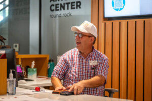 A man in a plaid shirt and white cap sits at a reception desk, smiling and gesturing. Behind him is a sign reading ARTS CENTER round rock. Office supplies and a computer are on the desk.