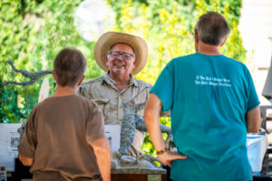 A man in a straw hat smiles while talking to two people at an outdoor event. Trees and informational signs are visible in the background.