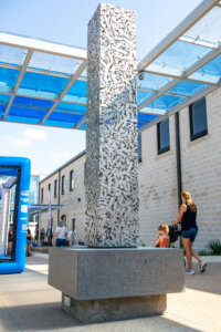 A tall, rectangular metal sculpture with abstract cut-out patterns sits on a stone base outdoors. People, including a woman and child, walk nearby under a blue pergola.