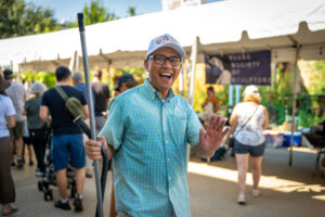 A smiling man in glasses and a cap waves while holding a stick at an outdoor event with a white tent and several people in the background.