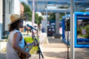 A musician wearing a large hat and sunglasses plays electric guitar and sings into a microphone outdoors, with people and booths visible in the background.