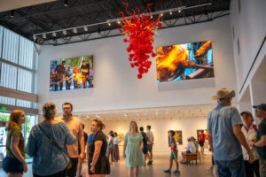 People gather inside a brightly lit art gallery with colorful glass sculptures and large screens showing glassblowing. A dramatic red glasswork hangs from the ceiling. Some people are chatting, while others admire the artwork.