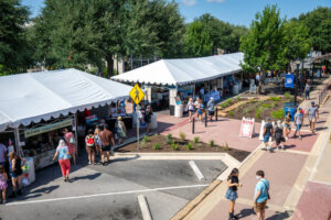 People browse vendor booths under white tents at an outdoor market on a sunny day. Shoppers walk along a paved path lined with greenery and trees.