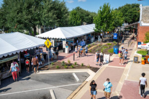 A street fair with white tents, vendors, and people walking around on a sunny day. There are trees, signs, and a mix of grass and paved areas along the street.