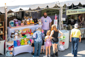 A group of people, including children, browse colorful crocheted stuffed animals at an outdoor market booth under a white tent. The vendor stands behind the display smiling. Other booths and shoppers are visible in the background.
