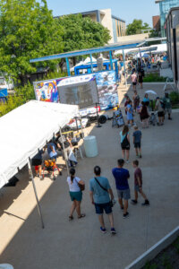 People walk and gather near a white tent and colorful trucks at an outdoor event on a sunny day, with trees and buildings in the background.
