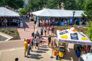 People gather at an outdoor festival with white tents for vendors, trees for shade, and a prominent Minute Maid tent in the foreground. Some visitors browse booths, while others walk along the paved paths.