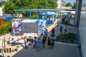 People gather around a race car exhibit with colorful artwork and charts, outdoors on a sunny day. Trees, tents, and a blue structure provide shade while visitors explore the area.