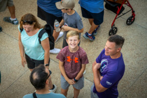 A smiling boy looks up at the camera, surrounded by adults and another child. People are gathered on a paved surface, some holding water bottles, with a red walker visible in the background.