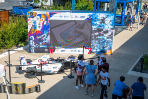 People gather around a white race car and a large screen displaying AI and robotics themes at an outdoor event. Colorful banners and a podium with 1, 2, 3 markings are visible. Some people take photos.