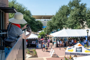 A person in a sunhat and red scarf takes a photo from a balcony overlooking a busy outdoor market with white tents, people walking, and trees under a clear blue sky.
