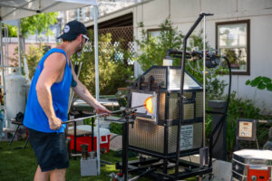 A man in a blue tank top shapes molten glass with a rod, standing beside a glassblowing furnace outdoors on grass, under a canopy.