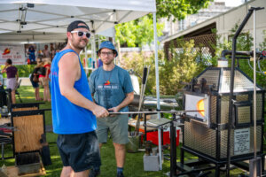 Two men demonstrate glassblowing outdoors under a tent. One man shapes molten glass on a rod, while the other smiles at the camera. Equipment and a furnace are visible, with onlookers and greenery in the background.