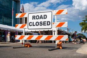 A large Road Closed sign blocks a street; people walk near tents at an outdoor event on a sunny day with a blue sky and scattered clouds.