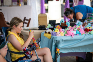A woman in a yellow shirt sits and crochets beside a table displaying various colorful crocheted stuffed animals at an outdoor market. Another person examines items on the table.