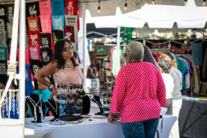 A woman stands behind a jewelry booth at an outdoor market, speaking to a customer wearing a red polka-dot shirt; colorful shirts hang in the background.