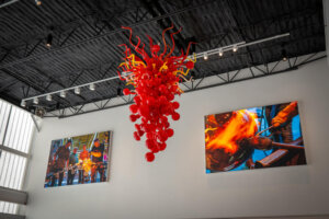 A vibrant red and yellow glass sculpture hangs from the ceiling of a modern gallery, with two large photos on the walls showing glassblowing in action.