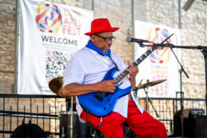A musician in a red hat and red pants plays a bright blue electric guitar on an outdoor stage during an arts festival.