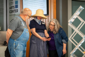 Three adults stand together indoors, looking at a smartphone. One wears denim overalls, another a wide-brimmed hat and scarf, and the third has long gray hair and glasses. They appear engaged with the phones screen.