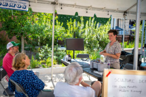 A person stands under a tent giving a demonstration to three seated people. A TV and supplies are on the table. Trees and buildings are visible in the background. A sign reading Sculpture Demo is in the foreground.