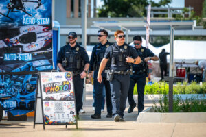 Four uniformed police officers walk on a sidewalk at an outdoor event, passing a STEM-themed sign with Take Your F4 Photo printed on it. Tents and trees are visible in the background.