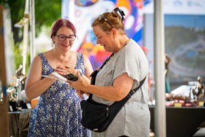 Two women stand and smile together at an outdoor market stall. One woman in a patterned dress holds her hands to her chest, while the other in a gray shirt writes on a pad. Colorful art and sculptures are visible in the background.