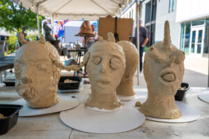 Three unfinished clay sculptures of heads, each with distinct features like horns and a unicorn horn, displayed on a table outdoors under a tent at an art event.