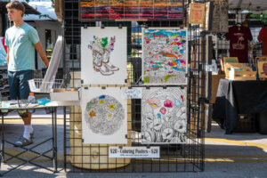 A display rack at an outdoor market holding four detailed coloring posters, some partially colored. A sign reads Coloring Posters - $20. A person stands nearby, and other vendor booths are in the background.