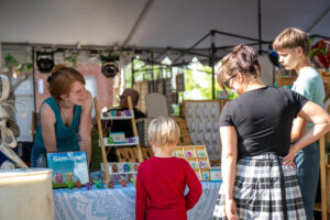 A vendor smiles and talks with a woman and two children at an outdoor market booth displaying colorful handmade jewelry and small figurines.