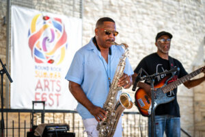 A man in sunglasses plays a saxophone onstage while another man plays bass guitar behind him at the Round Rock Summer Arts Fest.