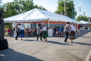 Six dancers in traditional attire perform on a street in front of a large white tent, while an audience watches seated under the tent on a sunny day.
