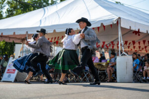 Four dancers in traditional costumes perform a folk dance outdoors, in front of a white tent with red flags, while an audience watches in the background on a sunny day.