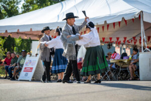 Four dancers in traditional clothing perform outside a white tent at a festival, with onlookers seated nearby. The dancers wear hats and skirts, and one raises an arm holding a handkerchief.