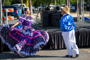 A woman in a purple traditional dress dances with a man in a blue shirt and straw hat outdoors near a stage, with musical equipment in the background.