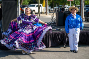 A woman in a colorful traditional dress dances beside a person in a blue shirt, white pants, and a straw hat on an outdoor stage with instruments and cars in the background.