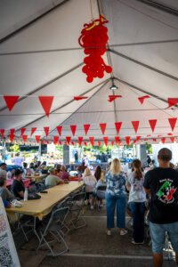 People sit at tables under a large white tent decorated with red pennant flags and a red balloon arrangement. Some people are standing, and a stage is visible in the background.