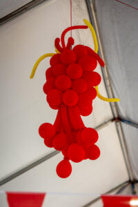 A cluster of red balloons shaped like a bunch of grapes hangs from the ceiling, with a few long yellow balloons at the top, inside a tent.