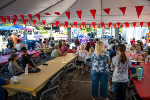 People sit under a tent decorated with red flags, watching a performer on stage. Tables are covered with yellow and red cloths, and the event takes place outdoors on a sunny day.