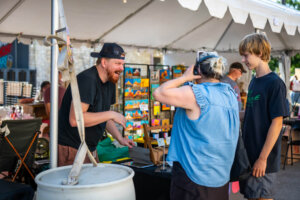 A vendor at an outdoor market booth smiles and talks with a woman and a boy standing nearby. Colorful art pieces are displayed in the background under a white canopy tent.