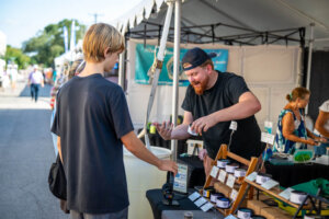 A vendor with a beard hands a product to a customer at an outdoor market stall. Other booths and people are visible in the background under a white tent.